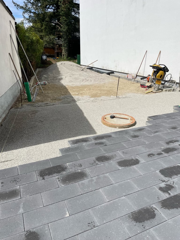 Residential patio under construction with gray paving stones, sandy work area, equipment, and building entrance visible