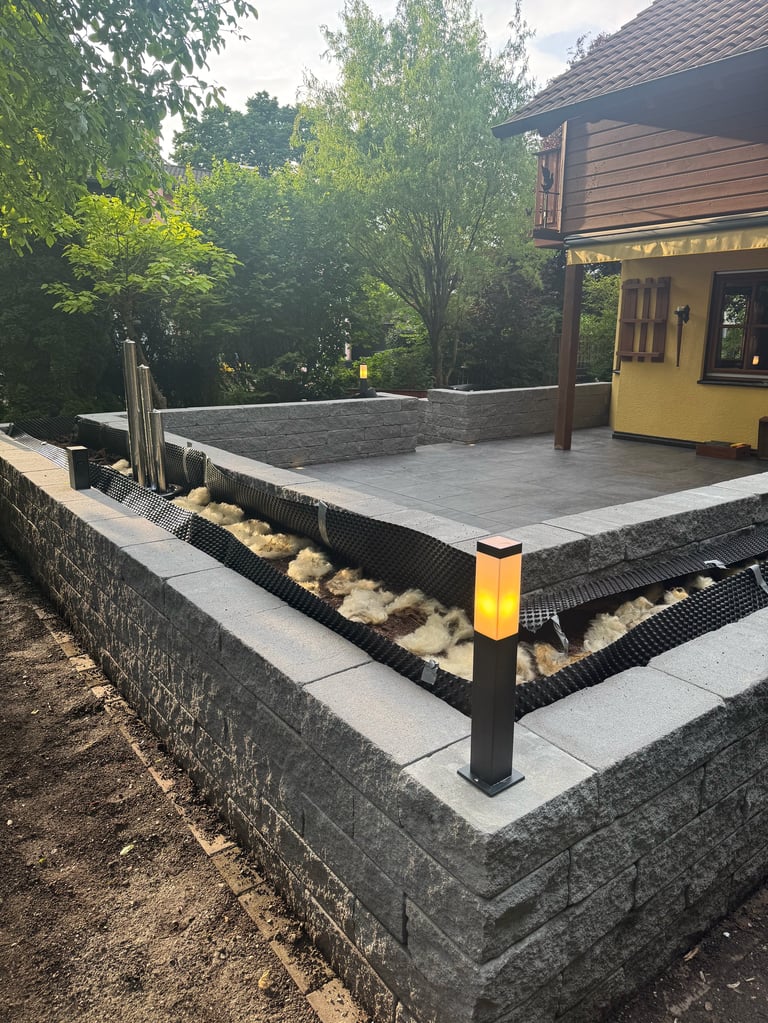 Modern residential patio with gray pavers, stone retaining walls, black bollard, and yellow-sided house surrounded by green trees