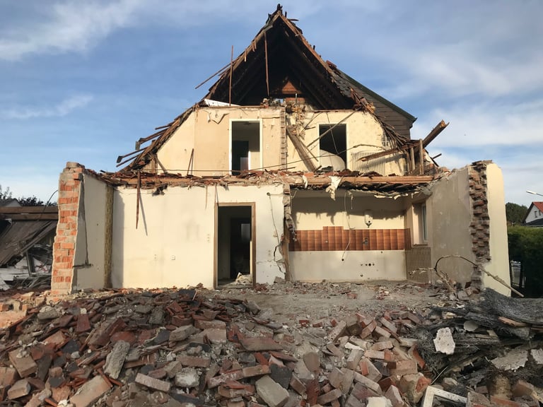 Severely damaged two-story house with collapsed roof and charred framework, surrounded by rubble and debris