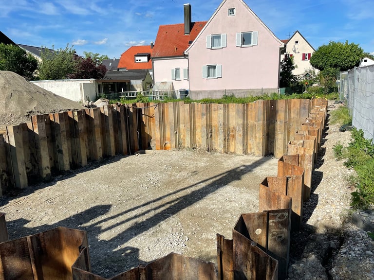 Residential construction site with wooden foundation piles and gravel, pink house visible in background under blue sky