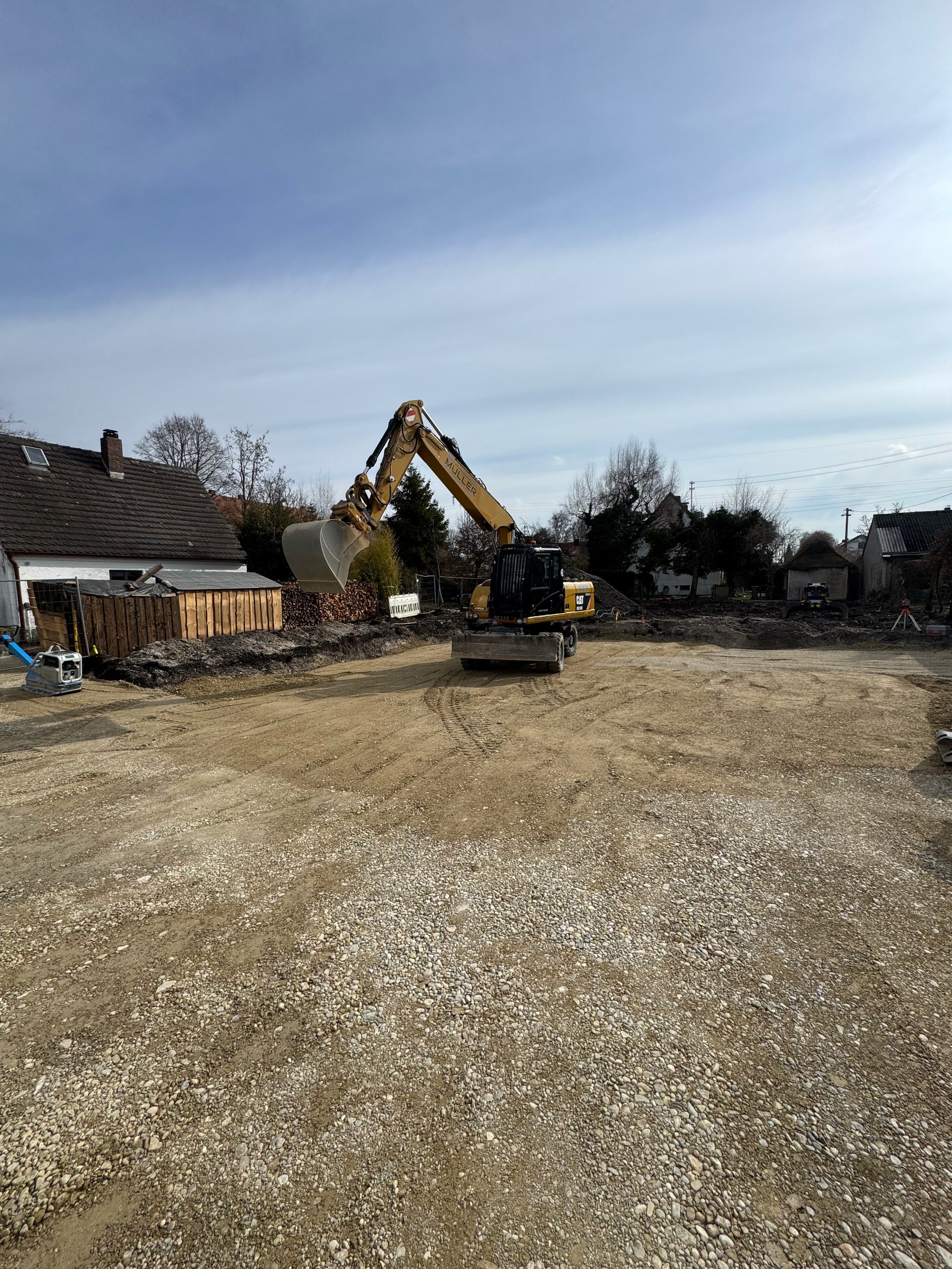 Yellow excavator on construction site with gravel ground, residential buildings, and blue sky in background