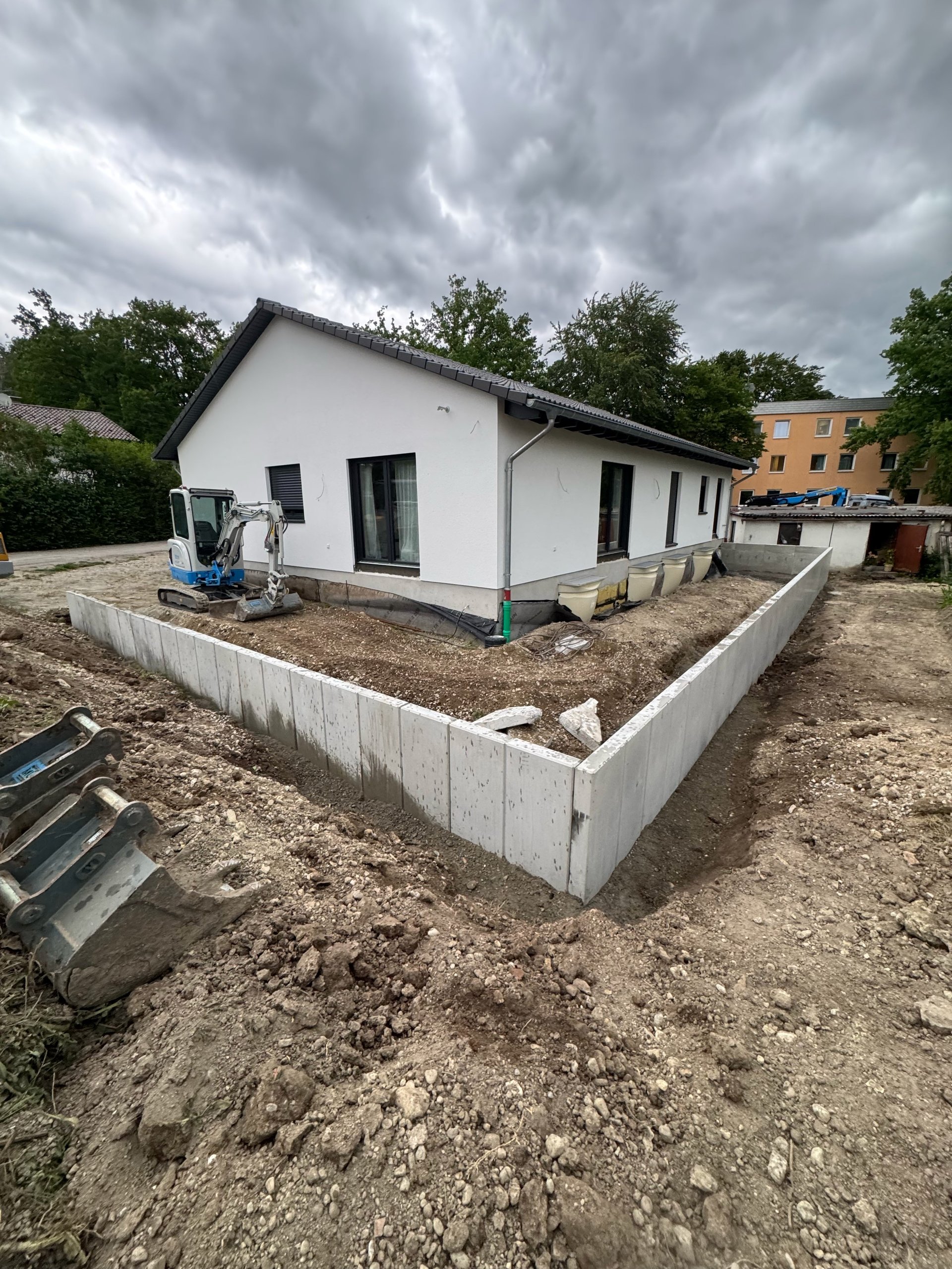 New white residential house under construction on concrete foundation with surrounding dirt, equipment, and grey stormy sky
