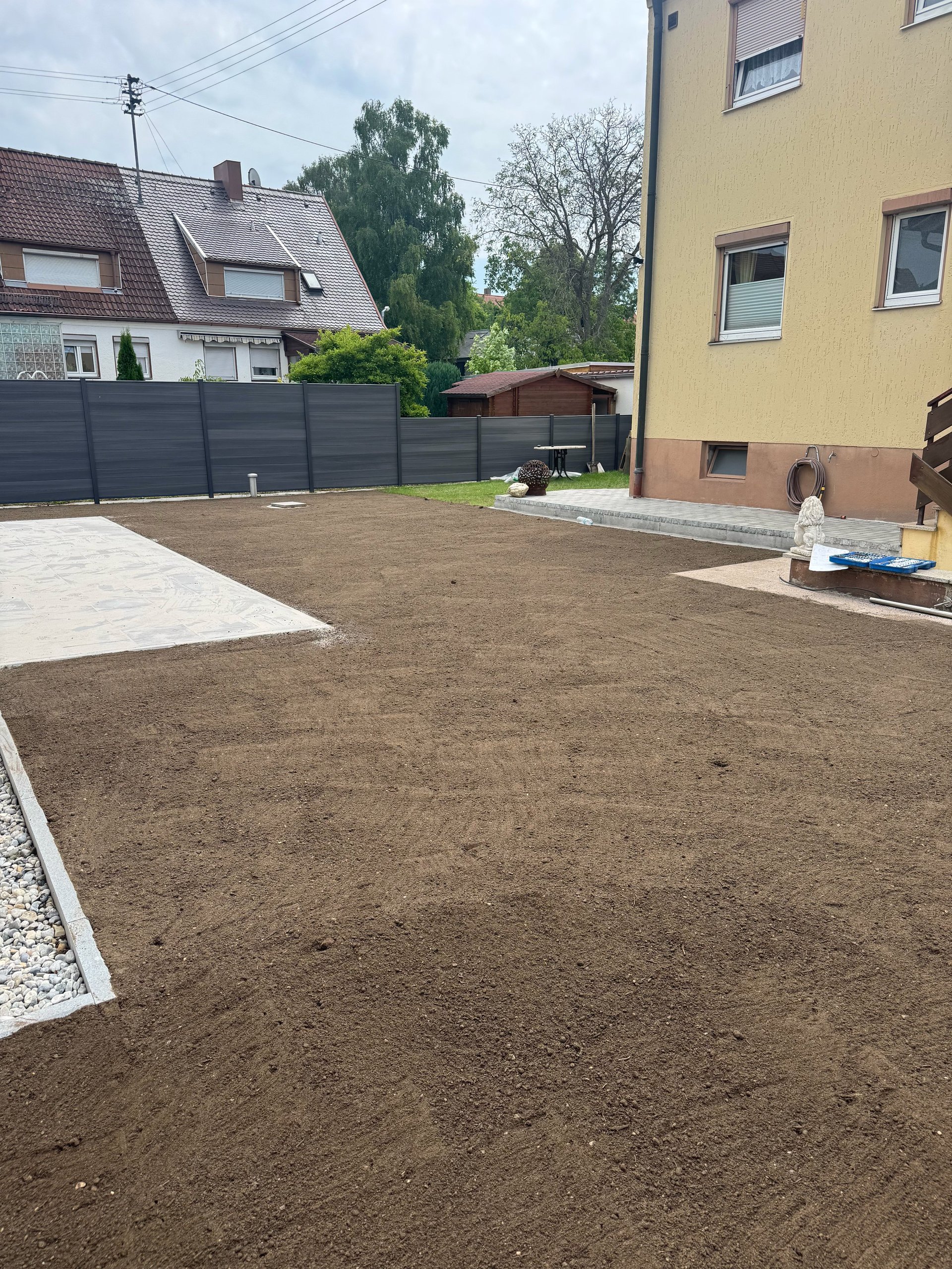 Backyard construction site with bare soil, yellow building, dark fence, and neighboring homes during landscaping preparation