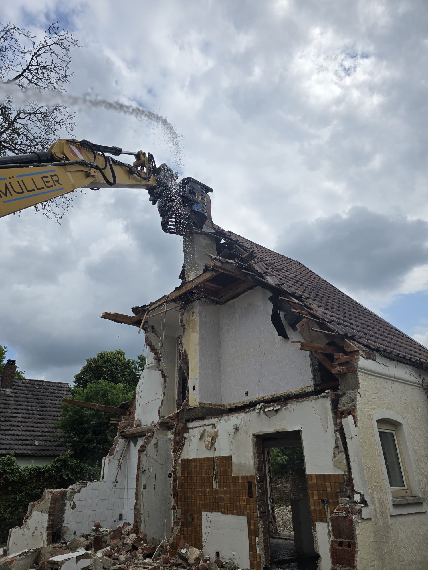 Excavator demolishing a white residential house, with structural debris visible and a cloudy sky overhead