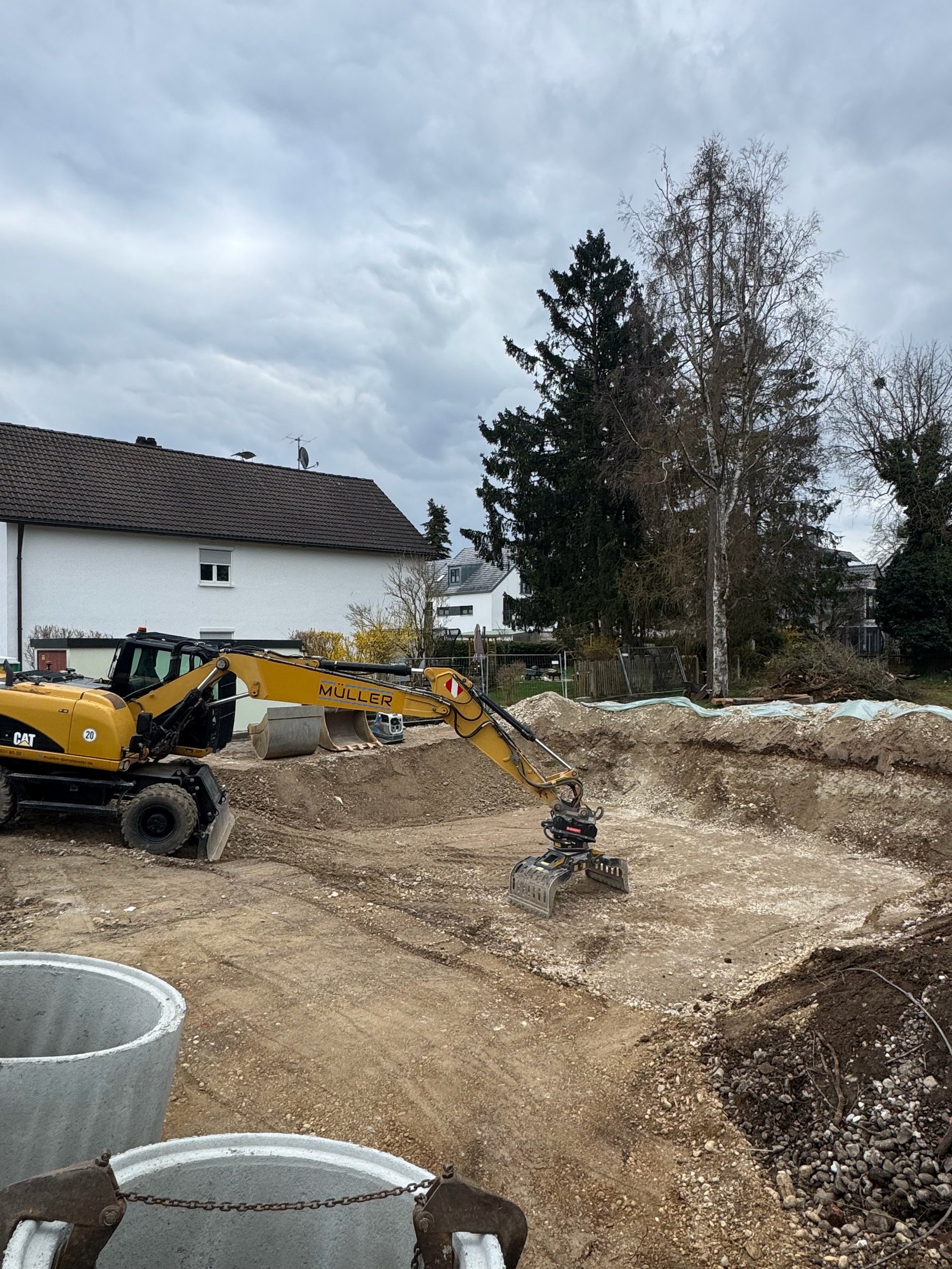 Yellow excavator on construction site with dirt mounds, white residential house and trees in background