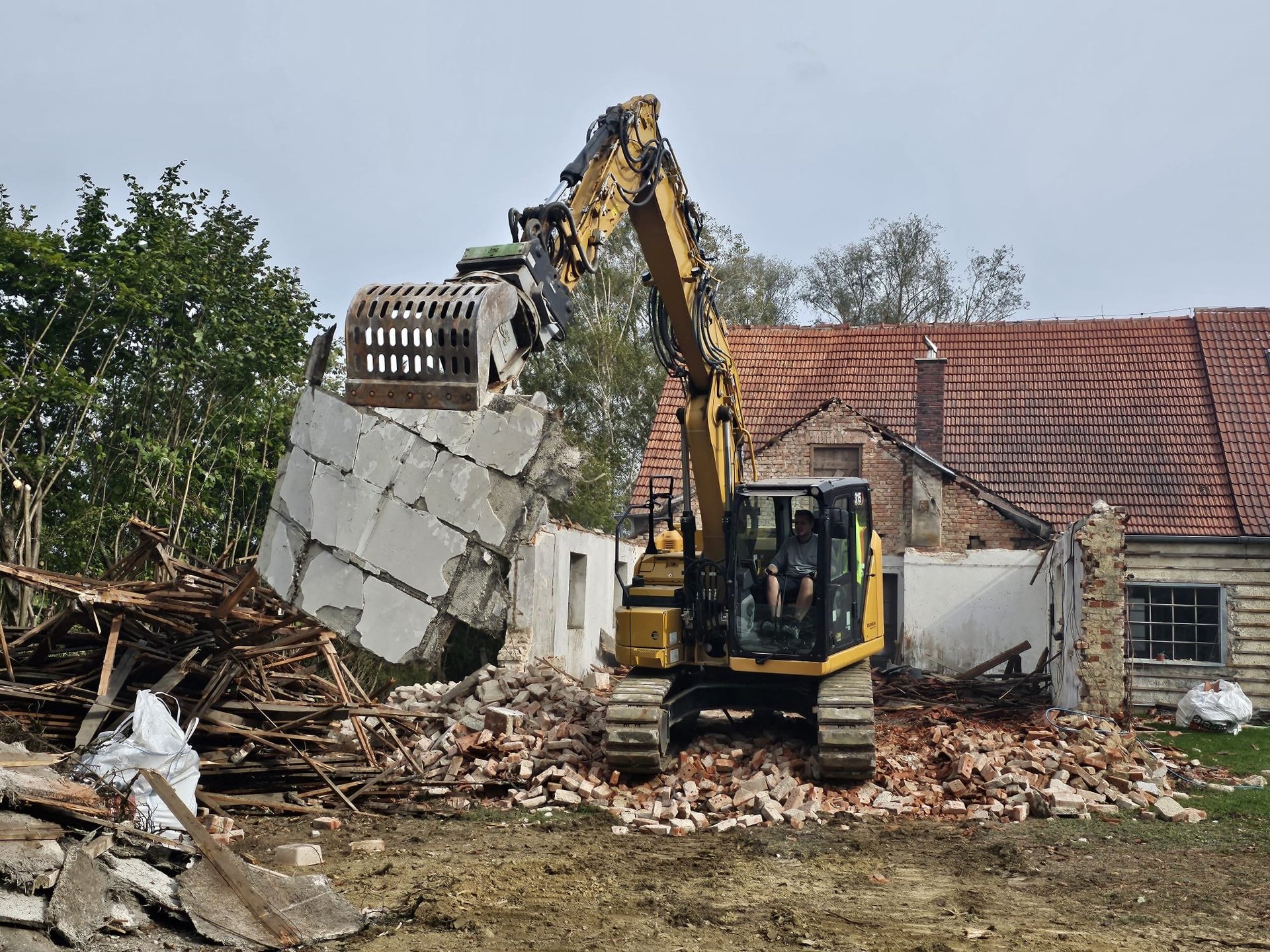 Excavator demolishing brick building with debris pile, red-roofed house visible in background