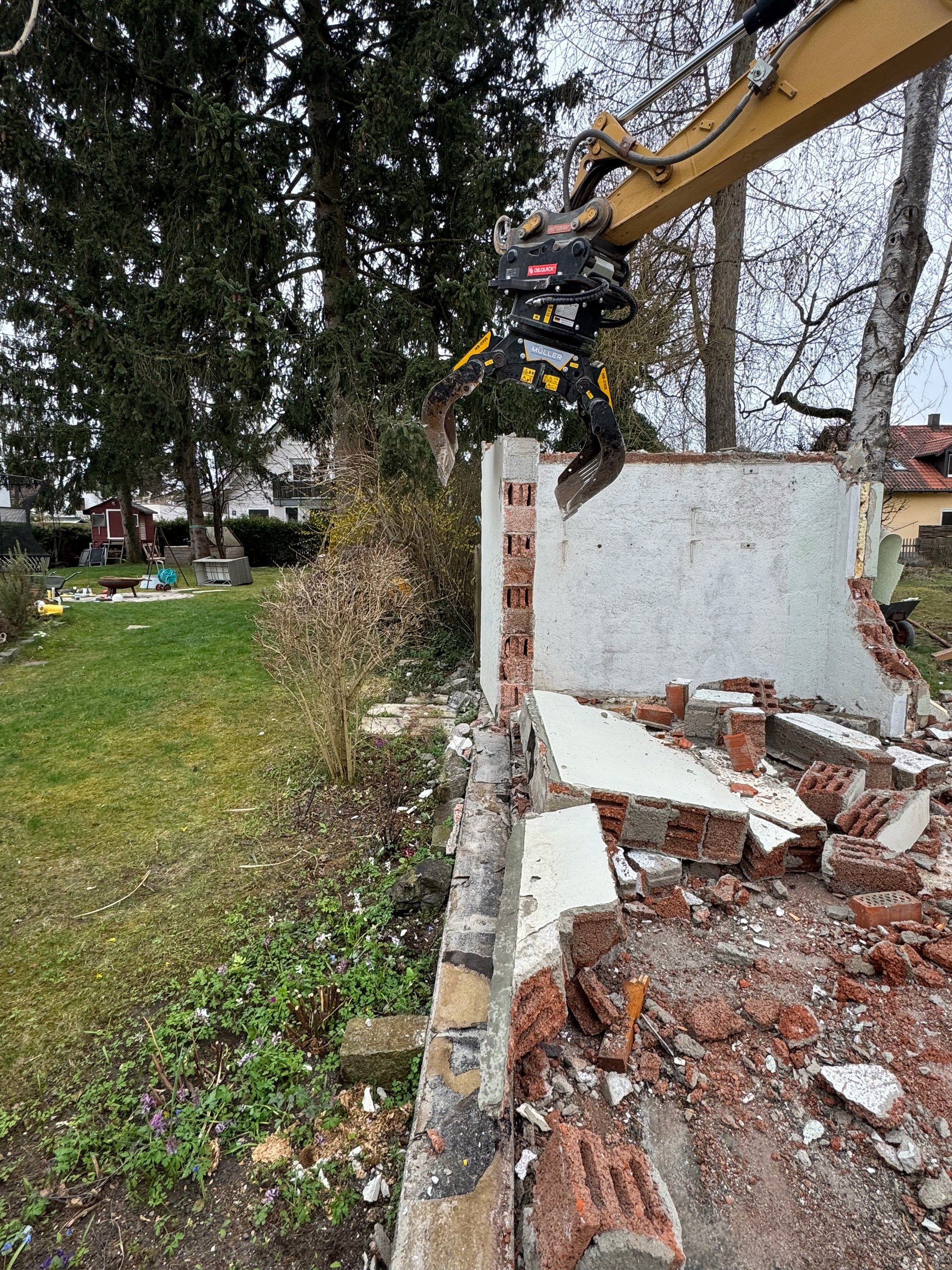 Excavator with grapple attachment demolishing white building structure next to residential property with trees