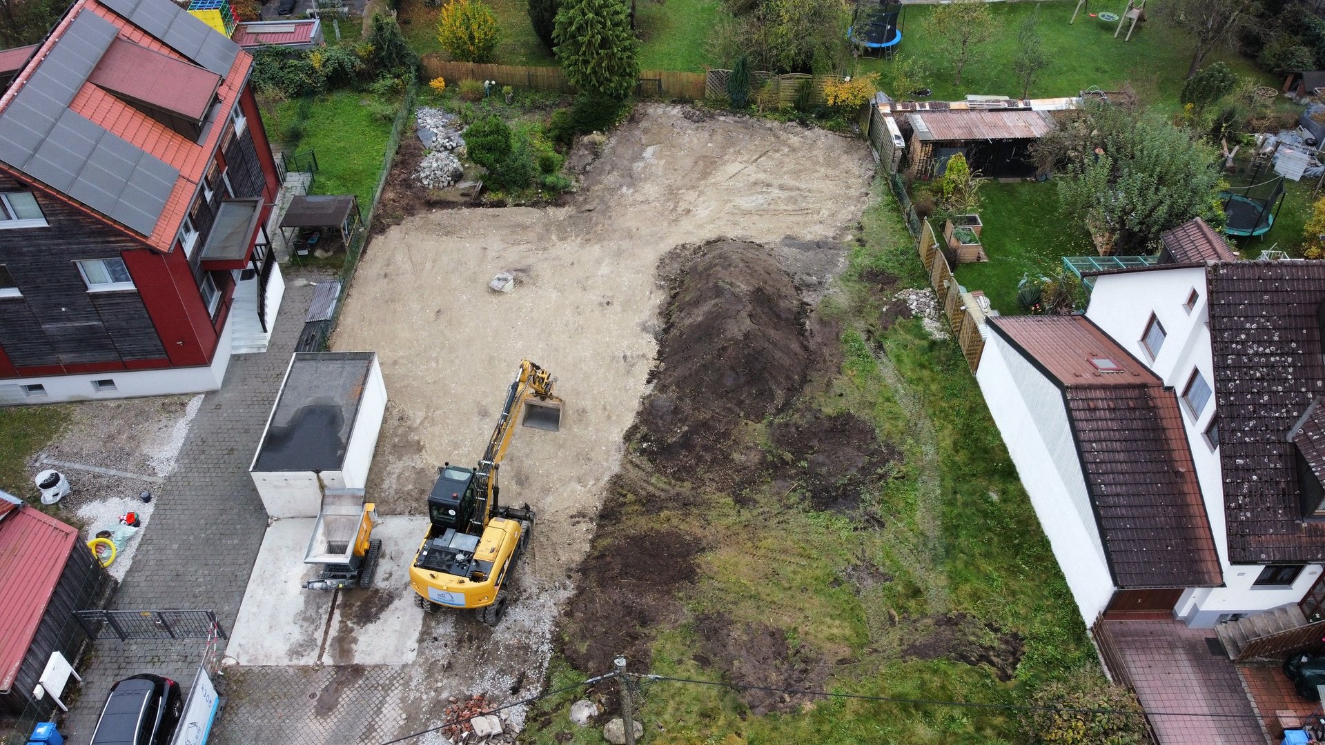 Aerial view of a construction site with excavator and machinery clearing land between residential buildings with red roofs