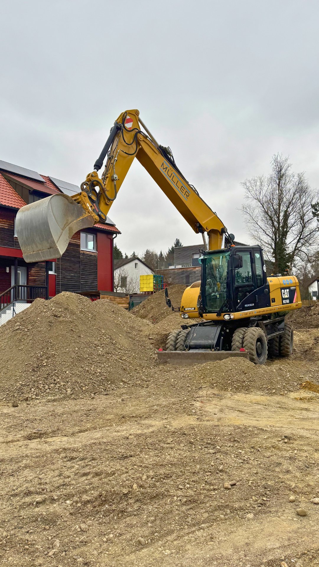 Yellow CAT wheeled excavator loading gravel onto a pile near residential buildings with red barn structures