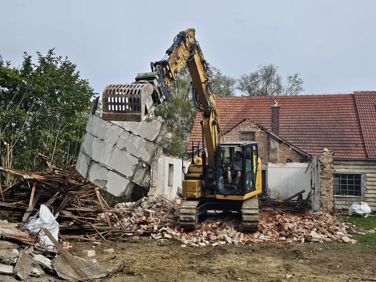 Yellow excavator demolishing concrete rubble at a construction site next to a brick house