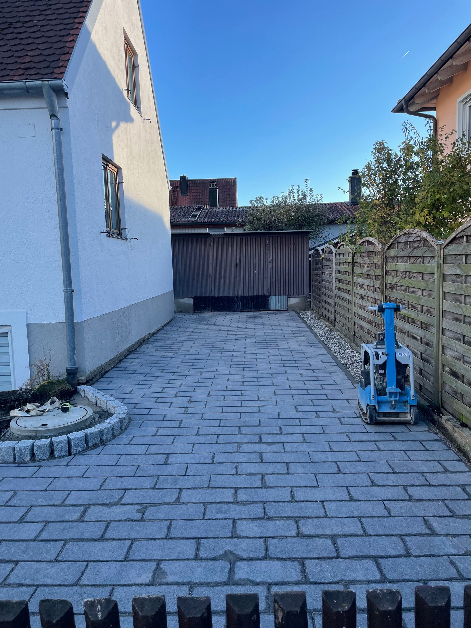 Paved driveway between white residential house and dark garage with wooden fence and blue equipment on right