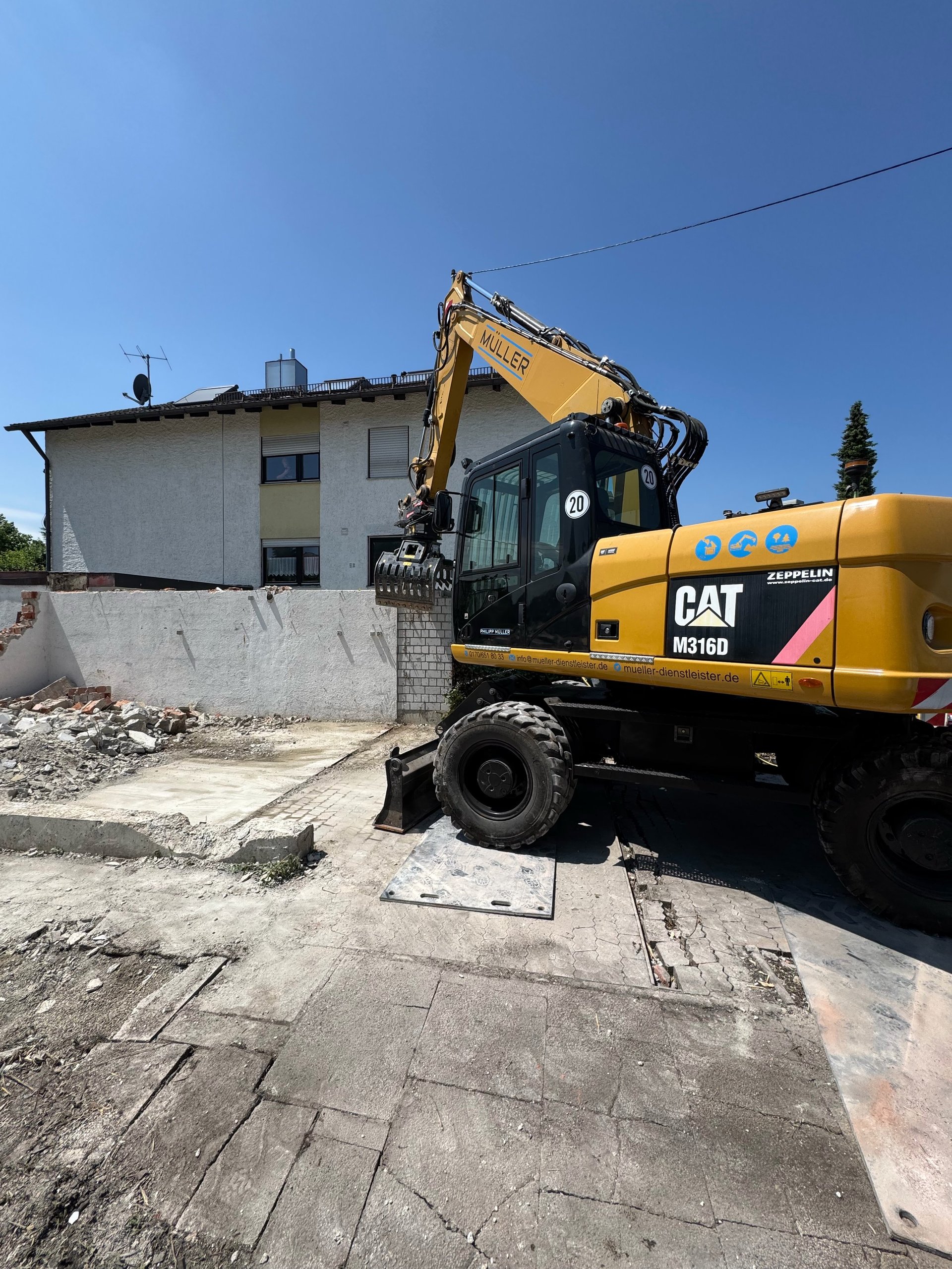 Yellow CAT M318 wheeled excavator demolishing a residential building on a sunny day with clear blue sky