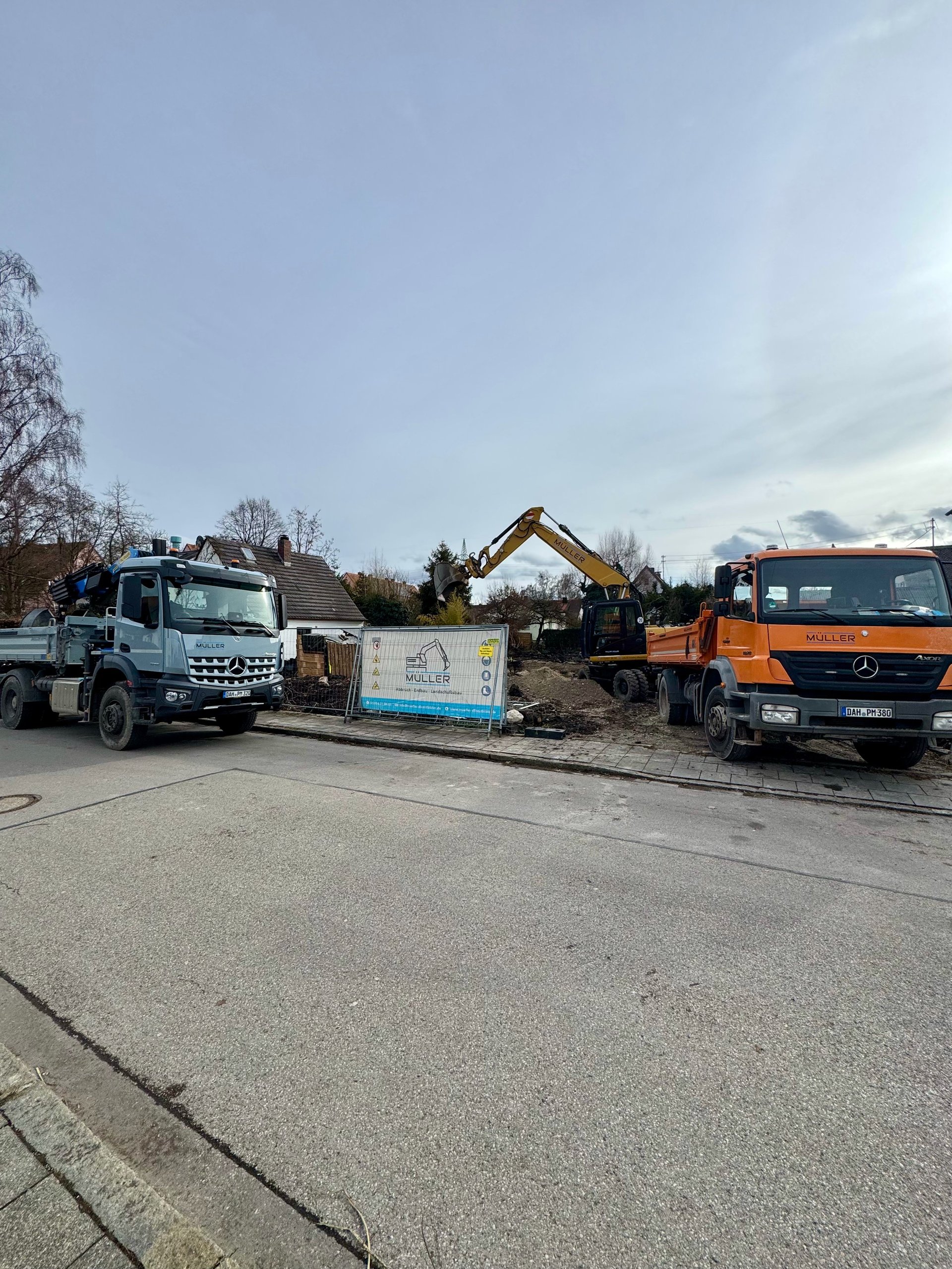 Construction site with a white truck on the left, an orange excavator in the center, and an orange Mercedes truck on the right, with bare trees and cloudy sky