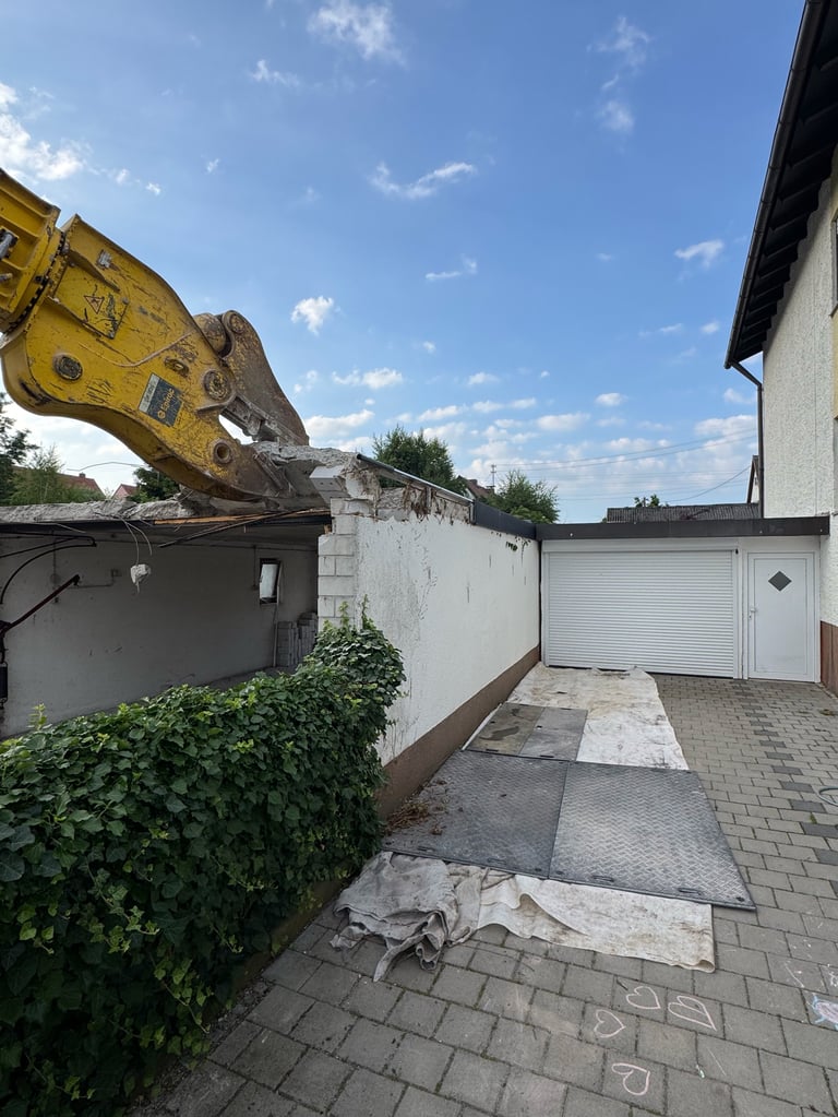 Yellow excavator with bucket extended over white industrial building in residential driveway with ivy and garage visible